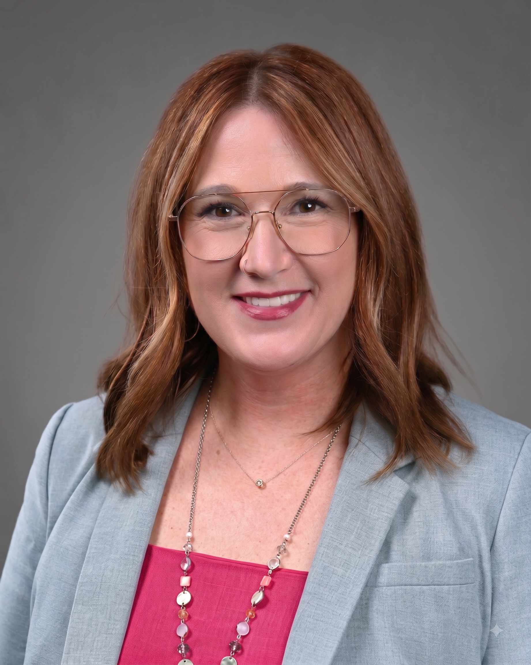 Smiling woman with a bluish grey suit jacket on with pink top.